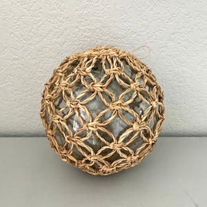 Puede incluir: Un flotador de vidrio redondo envuelto en una red tejida de fibra natural. El flotador es transparente y la red es de color marrón claro.