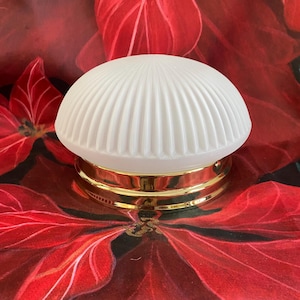 Op de afbeelding: Een witte, geribbelde glazen lamp met een goudkleurige basis. De lamp heeft een afgeronde, koepelvormige vorm en staat op een rode bloemenachtergrond. De lamp is waarschijnlijk voor een plafond of muur.