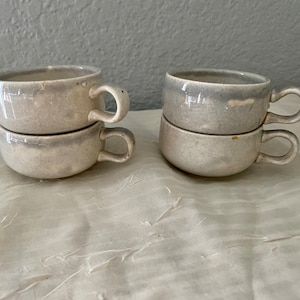 Könnte beinhalten: Vier kleine, handgefertigte Keramikbecher mit Henkeln, paarweise gestapelt. Die Becher sind hellgrau mit leicht strukturierter Oberfläche und dunklerem grauem Rand. Die Becher stehen auf einer weißen, strukturierten Oberfläche.