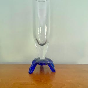 Può includere: Un vaso di vetro trasparente con una forma cilindrica svasata, appoggiato su una base blu vibrante dalla forma organica. Il vaso è esposto su una superficie di legno, mostrando il suo design elegante.