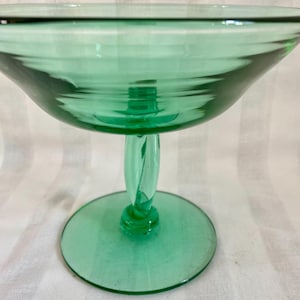 Può includere: Una coppetta da dessert in vetro verde con stelo. La coppetta ha una parte superiore ampia e poco profonda e una base arrotondata. Lo stelo ha un design a spirale. Il vetro ha una leggera tinta verde ed è trasparente.