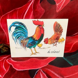 May include: A greeting card featuring a colorful rooster with a blue and green tail, red comb, and orange body. The card also depicts a hen and chicks, with the text "de colores!"
