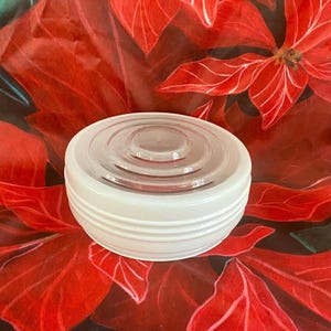 May include: A white, round, glass jar with a clear, ridged lid. The jar has a series of horizontal ridges around its base. The background features a red floral pattern.
