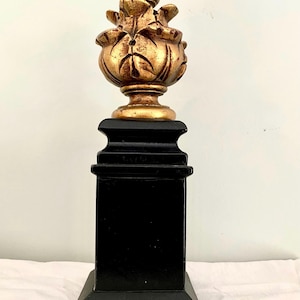 Puede incluir: Un adorno decorativo con una parte superior dorada y ornamentada y una base negra. La parte superior tiene un diseño floral detallado. La base es un pedestal cuadrado y escalonado. El adorno probablemente está hecho de madera o un material similar.