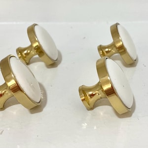 Puede incluir: Cuatro pomos de armario con acabado dorado e inserciones ovaladas blancas. Los pomos están dispuestos en grupo, mostrando su diseño y uso potencial en decoración del hogar o proyectos de mobiliario.