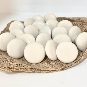 May include: A group of smooth, cream-coloured ceramic stones, displayed on a textured, tan hessian cloth. The stones are round and of varying sizes, creating a natural, organic look. The neutral colours and tactile textures suggest a decorative or craft theme.