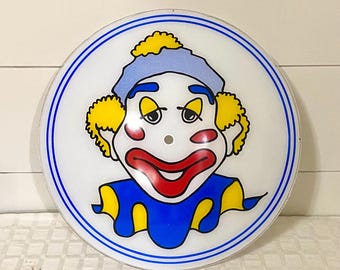 以下が含まれることがあります： カラフルなピエロのグラフィックが描かれた円形のガラス製照明カバー。ピエロは黄色い髪、青い帽子、赤い頬、大きな赤い口をしています。デザインは白い背景に青い円形の境界線で構成されています。