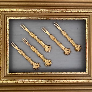 May include: Six gold-colored, decorative forks with intricate designs are displayed against a gray background within a gold-colored frame.