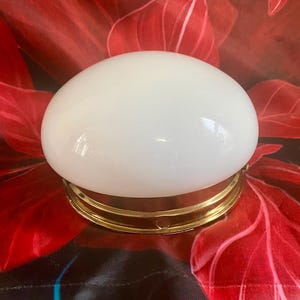 Könnte beinhalten: Eine ovale, weiße Glasleuchte mit einem goldfarbenen Metallsockel. Die Leuchte ist vor einem roten floralen Hintergrund platziert. Die Leuchte ist ein dekoratives Element für die Inneneinrichtung.