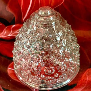 Pode incluir: Um objeto decorativo de vidro transparente com uma superfície texturizada em forma de bolhas. O objeto tem uma forma arredondada em forma de cúpula e está sobre um fundo floral vermelho. O vidro é transparente, permitindo a passagem da luz.