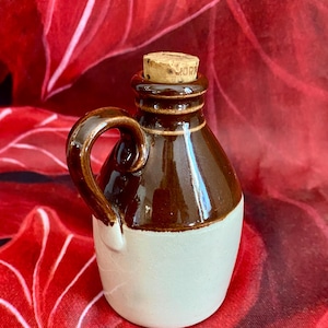 May include: A small, brown and white ceramic jug with a handle and a cork stopper. The jug has a rounded body and a dark brown top half, with a lighter brown handle. The bottom half is white. The cork stopper has the word "MORR" printed on it.