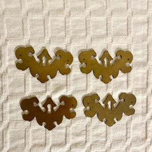May include: Four antique brass drawer pulls with a decorative, symmetrical design. Each pull features a keyhole and a stylized, ornate shape with curved edges and a warm, aged patina. These vintage hardware pieces are ideal for furniture restoration.