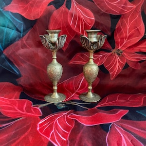 May include: Two brass candle holders with a floral design. The candle holders have a tulip-shaped top and a detailed, ornate base. The background is a red and black floral pattern, creating a festive and decorative display.