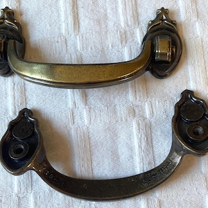 Könnte beinhalten: Zwei antike Schubladengriffe aus Messing mit aufwendigen Details. Die Griffe haben ein gebogenes Griffdesign und dekorative Endstücke. Einer der Griffe hat eine hellere Messingfarbe, der andere ist ein dunkleres, gealtertes Messing. Sie sind wahrscheinlich für die Möbelrestaurierung.