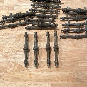 May include: A collection of antique brass cabinet pulls and handles. The pulls have a detailed, ornate design with a dark, aged finish. The handles are vertical with a similar design. The hardware is arranged on a light wood surface.