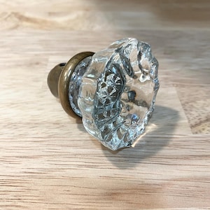May include: A clear, faceted glass cabinet knob with a circular, multi-sided design. The knob features a reflective, textured surface and is attached to a circular, antique-toned brass metal base that connects to a cylindrical mounting post.