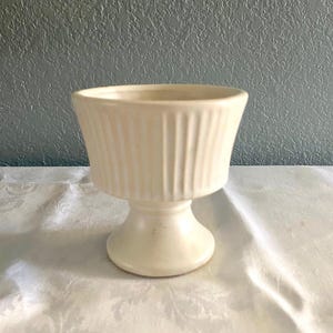 May include: Cream-colored ceramic pedestal bowl with vertical ribbed detailing. The bowl has a wide, open top and a rounded base, set on a short pedestal. The background is a textured, light blue wall.
