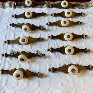 Puede incluir: Un conjunto de tiradores de armario antiguos con acabado de latón y pomos de cerámica color crema. Los tiradores tienen un diseño decorativo y ornamentado y están dispuestos sobre una superficie blanca. Ideales para proyectos de restauración de muebles.