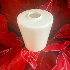 May include: A white, opaque glass lampshade with a slightly tapered cylindrical shape. The lampshade has a rounded top with a central opening. The background features a red floral pattern.