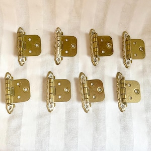 May include: Eight brass-colored cabinet hinges with decorative edges. Each hinge has a rectangular plate with three screw holes and a hinged section. The hinges are arranged on a striped white background.