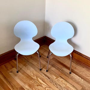 May include: Two light blue chairs with a unique, organic shape. The chairs have chrome legs and are set against a neutral wall and wooden floor. The chairs are a modern design.
