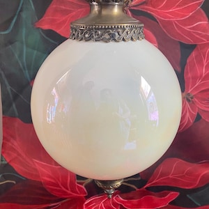 Könnte beinhalten: Eine Hängeleuchte im Vintage-Stil mit einer runden, cremefarbenen Glaskugel. Die Leuchte hat verzierte, messingfarbene Metalldetails an der Ober- und Unterseite. Der Hintergrund zeigt ein Blumenmuster mit roten, poinsettienartigen Blüten.