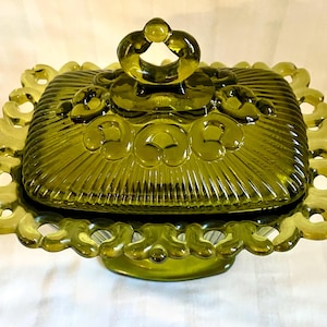 Pode incluir: Uma manteigueira de vidro verde-oliva com tampa. O prato retangular tem uma borda decorativa com vieiras e uma alça circular na tampa. O vidro tem uma textura canelada. Uma peça vintage.