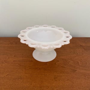 Puede incluir: Un cuenco de caramelos blanco vintage con pedestal. El cuenco tiene un cuenco redondo y abierto con un borde festoneado y una base circular. El cuenco está hecho de vidrio opaco y se encuentra sobre una superficie de madera.