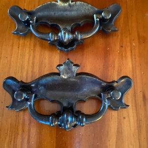 Może przedstawiać: Dwa antyczne uchwyty do szuflad z brązu z ozdobnymi detalami. Każdy uchwyt posiada zakrzywiony uchwyt, dekoracyjne centrum i elementy montażowe. Uchwyty są ustawione na tle usłojenia drewna.