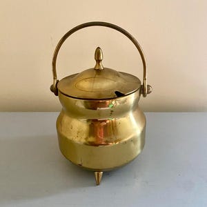 Puede incluir: Una pequeña olla de latón de color dorado con tapa y asa. La olla tiene tres patas puntiagudas y un cuerpo redondeado.
