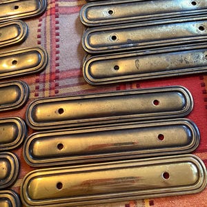 May include: A selection of antique brass drawer pulls. Each pull is rectangular with rounded ends and a single central hole for mounting. The pulls have a warm, aged patina, suggesting they are vintage or vintage-inspired.