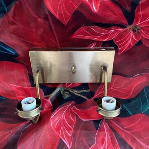 May include: A vintage brass wall sconce with two light sockets. The sconce has a rectangular backplate and curved arms holding the sockets. The background features a red floral pattern.
