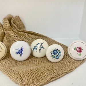 May include: Five decorative ceramic cabinet knobs with floral and bird designs. The knobs are white and cream-colored, with blue and pink floral patterns. They rest on a burlap sack.