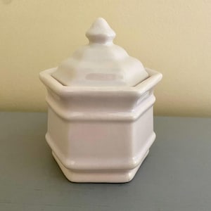 Peut inclure: Un pot hexagonal en céramique blanche avec un couvercle. Le couvercle a un sommet pointu et une finition lisse et brillante.