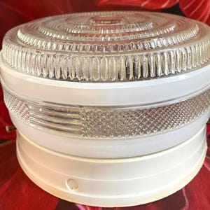 May include: A vintage ceiling light fitting with a clear glass shade and a white base. The glass shade has a ribbed design around the edges and a circular pattern on top. The white base has a rounded shape.
