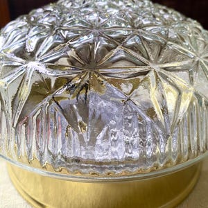 May include: A clear glass light fixture with a textured, geometric pattern dome. The base is a shiny gold color. The fixture is designed to be mounted on a ceiling and provides ambient lighting.