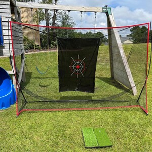 May include: A golf practice net with a black target and red trim, set up on a grassy lawn. A small green golf mat is in front of the net. A blue slide and wooden play structure are in the background.