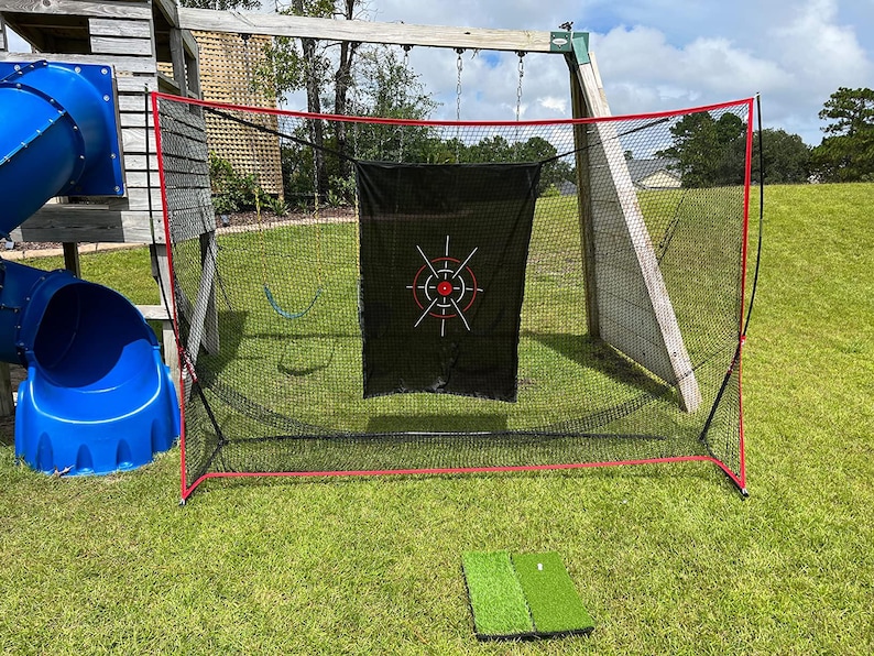 May include: A golf practice net with a black target and red trim, set up on a grassy lawn. A small green golf mat is in front of the net. A blue slide and wooden play structure are in the background.