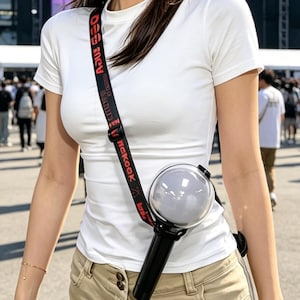 Op de afbeelding: Een vrouw in een wit T-shirt en een kaki broek lacht. Ze houdt een zwarte en transparante lichtstick vast met een rode "BOROK"-band. Op de achtergrond zijn een groot stadion en een menigte te zien.