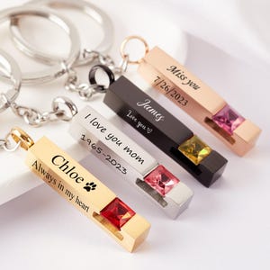 Puede incluir: Una colección de llaveros personalizados en oro, plata, negro y oro rosa. Cada colgante rectangular presenta una piedra preciosa y texto grabado, incluyendo nombres, fechas y frases como "Always in my heart" e "I love you mom."