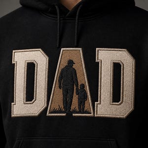 May include: Black hoodie with the word "DAD" embroidered in a tan, block letter style. The letter "A" features an embroidered image of a person and child walking together. The hoodie has a drawstring hood.