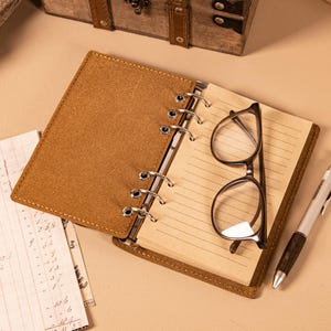 May include: Open brown leather journal with lined pages, a pair of eyeglasses, and a pen. The journal is open, revealing the interior pages. The eyeglasses are black-framed. The pen has a silver tip and a wooden handle.