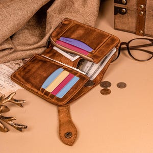 Puede incluir: Cartera de cuero marrón abierta con múltiples ranuras para tarjetas y un compartimento con cremallera. La cartera contiene efectivo y tarjetas de colores. Un cierre a presión asegura la cartera. Hay monedas esparcidas cerca.