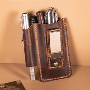 Könnte beinhalten: Ein brauner Leder-Multitool-Halter mit Metallclip, der einen Stift, ein Messer und andere Werkzeuge enthält. Der Halter hat sichtbare Nähte und ein abgenutztes Aussehen, was auf Haltbarkeit und Nutzung hindeutet.