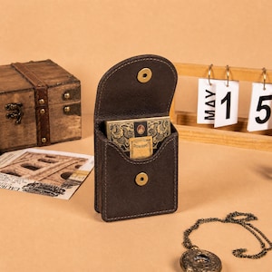 Peut inclure: Une pochette en cuir marron foncé avec un rabat et un bouton-pression doré. La pochette contient un jeu de cartes à jouer avec des motifs dorés et noirs ornés et une petite étiquette rectangulaire avec le texte "THE KORN" imprimé dessus.
