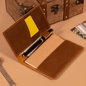 Op de afbeelding: Open bruin leren notitieboek met een gele kaart in de zak, een zwarte pen en een notitieblok. Het notitieboek is open en onthult de binnenkant. De achtergrond omvat een houten kist en een foto.