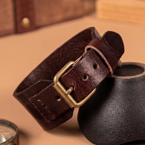 May include: A dark brown leather cuff bracelet with a brass buckle. The bracelet is thick and has a textured surface. The buckle is rectangular and has a classic design. The bracelet is approximately 5 cm wide.