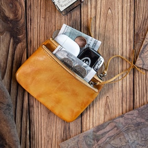 May include: A tan leather zipped pouch containing a white wireless earbud case, a black rectangular electronic device, and several coins. The pouch features a thin leather pull tab attached to the metal zip, showcasing a smooth, natural texture.