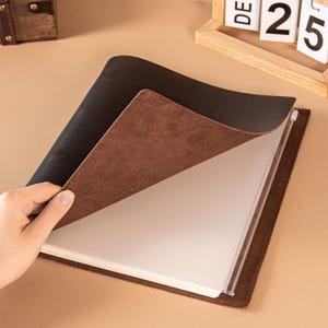 Puede incluir: Un diario de cuero marrón con una cubierta interior marrón oscuro está abierto, revelando fundas de plástico transparente para mostrar fotos o documentos. El diario está sobre una superficie marrón claro.