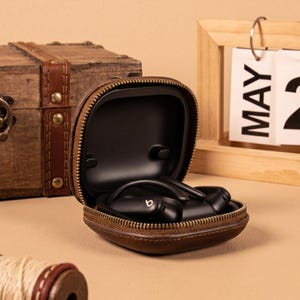 Könnte beinhalten: Ein Paar schwarze kabellose Ohrhörer mit Ohrbügeln und dem Beats-Logo, aufbewahrt in einer braunen Leder-Reißverschlusstasche. Das Etui ist geöffnet und zeigt ein schwarzes Innenfutter, das die Ohrhörer sicher an ihrem Platz hält.